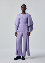 A person stands against a plain background wearing the MOTION BLUR lavender pleated jersey top with wide, layered sleeves and matching wide-legged pants with front slits by HOMME PLISSÉ ISSEY MIYAKE, styled with short hair and black shoes.