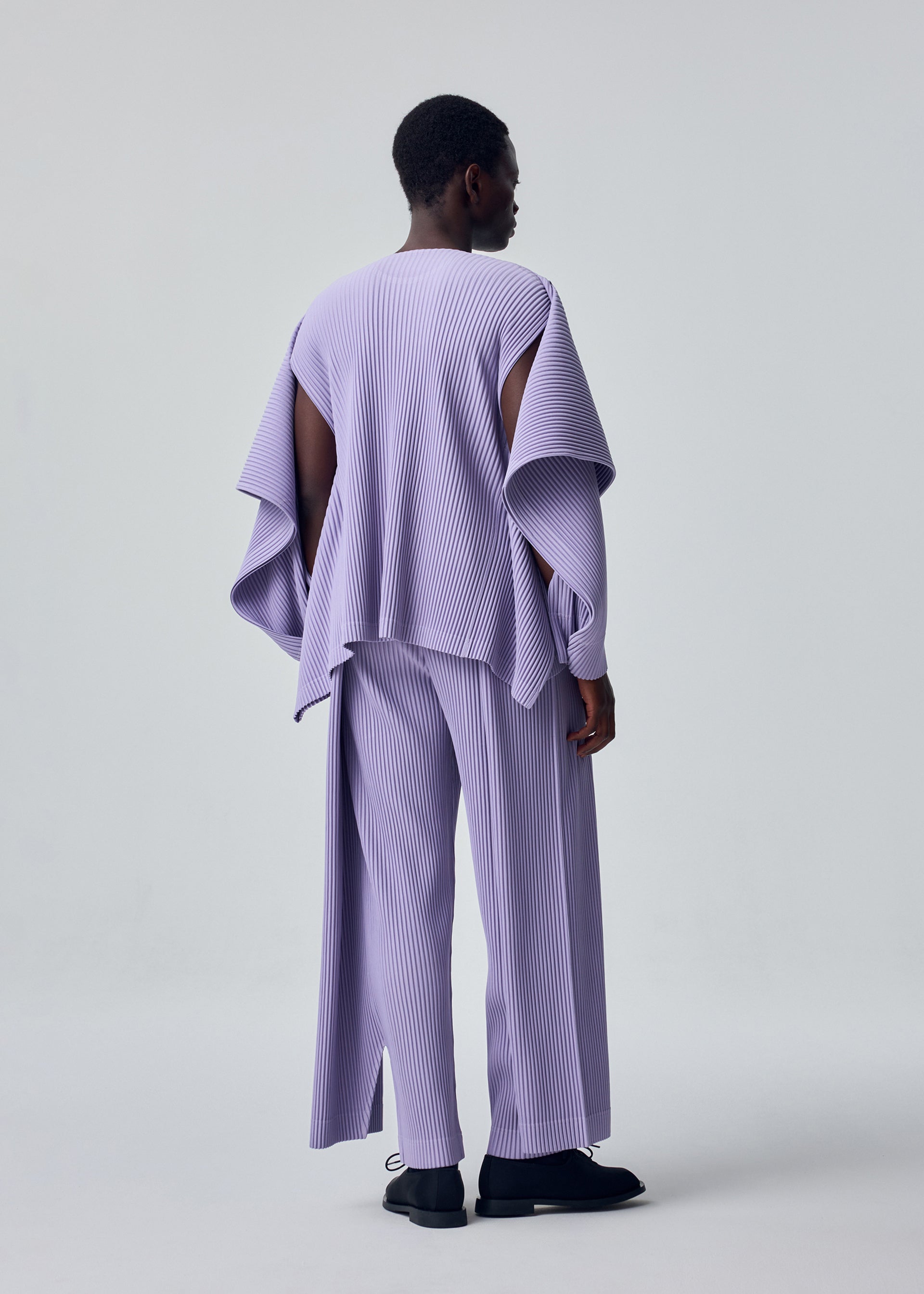 Zoomed view A person faces away, wearing HOMME PLISSÉ ISSEY MIYAKE's MOTION BLUR—a loose, pleated jersey top with wide sleeves and matching pants. The plain light background highlights its modern texture and versatile styling options.