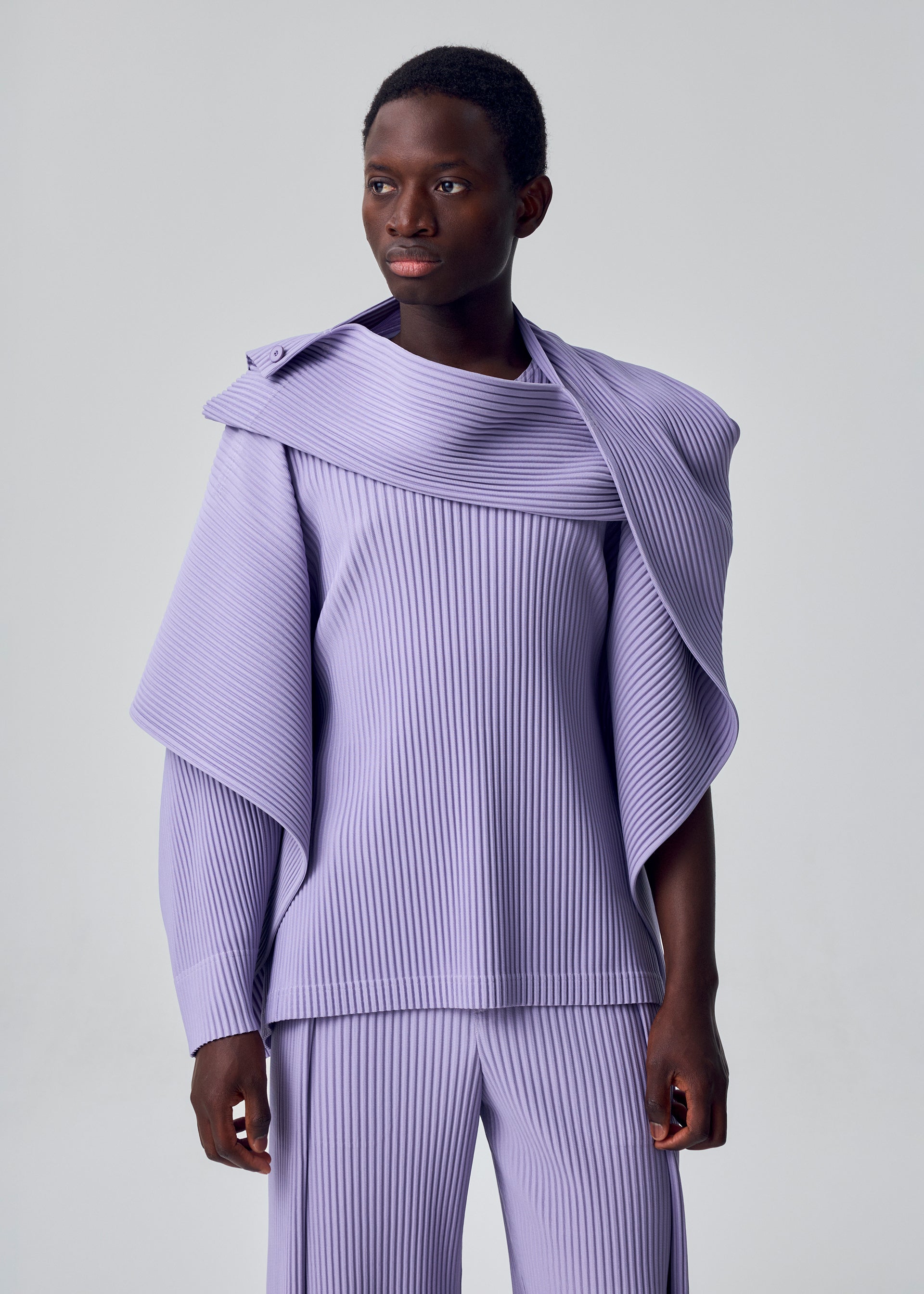 Zoomed view A person showcases the versatile MOTION BLUR set from HOMME PLISSÉ ISSEY MIYAKE—a stylish light purple pleated jersey top with an asymmetrical drape and matching pants—against a plain, light gray background.