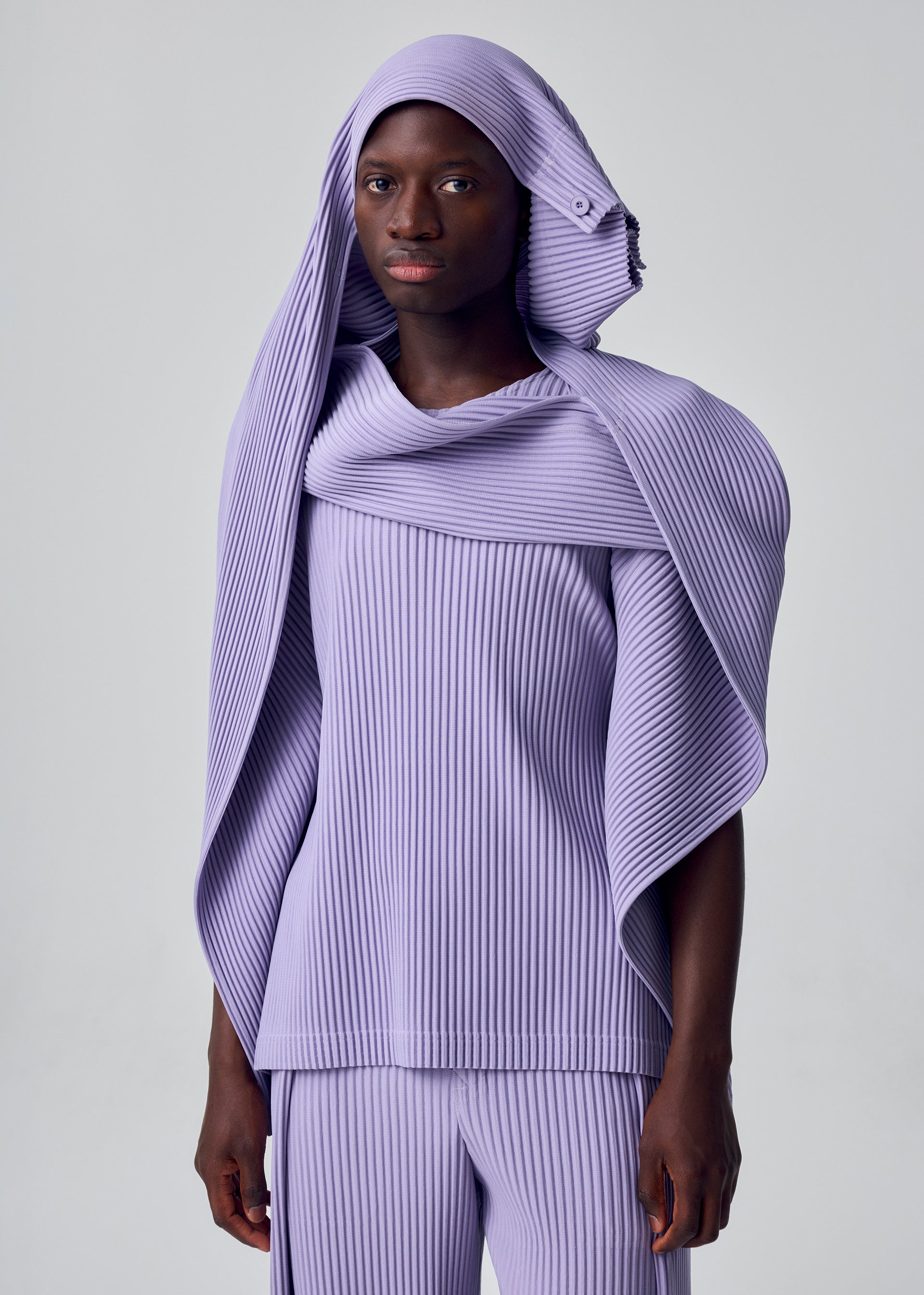 Zoomed view A person wears the HOMME PLISSÉ ISSEY MIYAKE MOTION BLUR set—a ribbed lavender outfit with matching pants and a pleated jersey top featuring a versatile draped hood—posed against a plain light background.