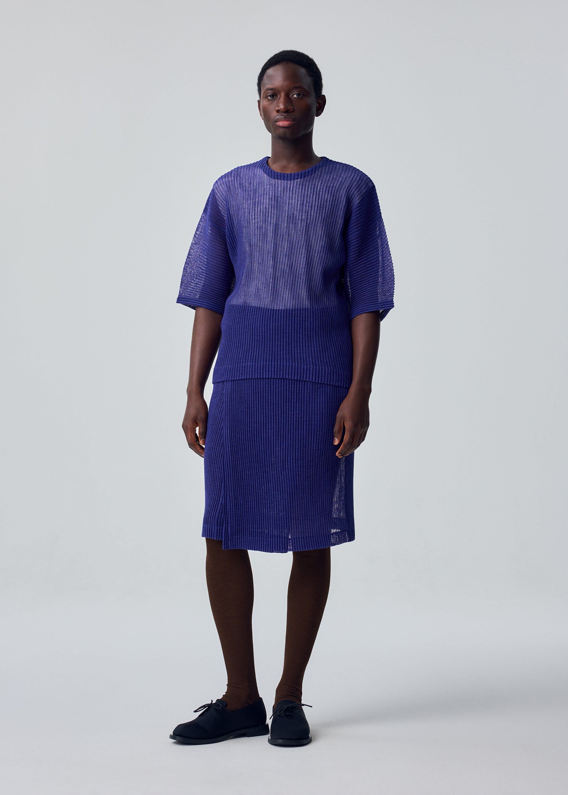 Zoomed view A person faces the camera with a neutral expression, wearing the RUSTIC MESH sheer mesh set by HOMME PLISSÉ ISSEY MIYAKE, paired with brown tights and black shoes against a plain light background.
