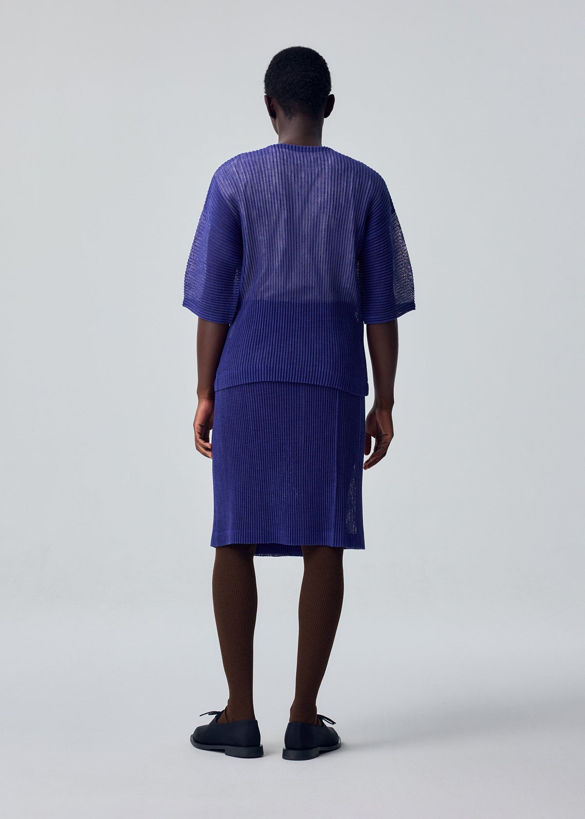 A person faces away, wearing HOMME PLISSÉ ISSEY MIYAKE's RUSTIC MESH—a loose, pleated purple top with a matching knee-length skirt—paired with brown tights and black shoes. The background is plain and light-colored. view 6 of 6