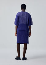 A person faces away, wearing HOMME PLISSÉ ISSEY MIYAKE's RUSTIC MESH—a loose, pleated purple top with a matching knee-length skirt—paired with brown tights and black shoes. The background is plain and light-colored.