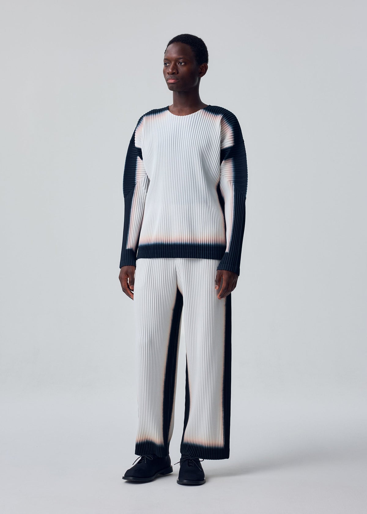 A person wears the FRAMEWORK : CHUSEN top by HOMME PLISSÉ ISSEY MIYAKE—white, pleated, with black gradient edges—paired with black shoes against a plain light background. view 4 of 5