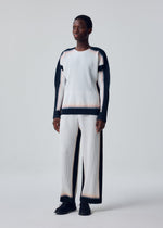 A person wears the FRAMEWORK : CHUSEN top by HOMME PLISSÉ ISSEY MIYAKE—white, pleated, with black gradient edges—paired with black shoes against a plain light background.