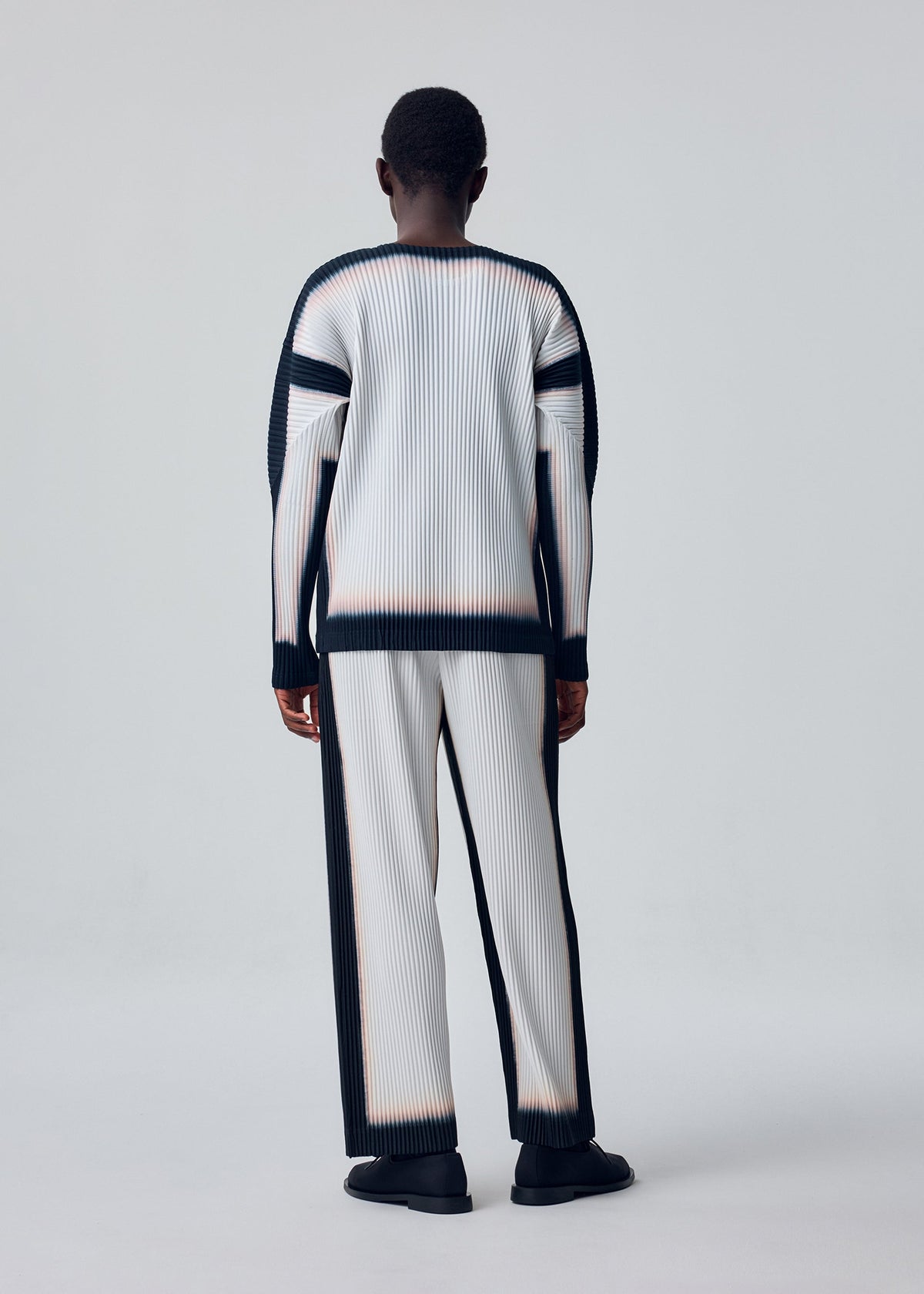 A person faces away, wearing the FRAMEWORK : CHUSEN top by HOMME PLISSÉ ISSEY MIYAKE—white, pleated, long-sleeved with black trim—paired with black shoes against a plain light background. view 5 of 5