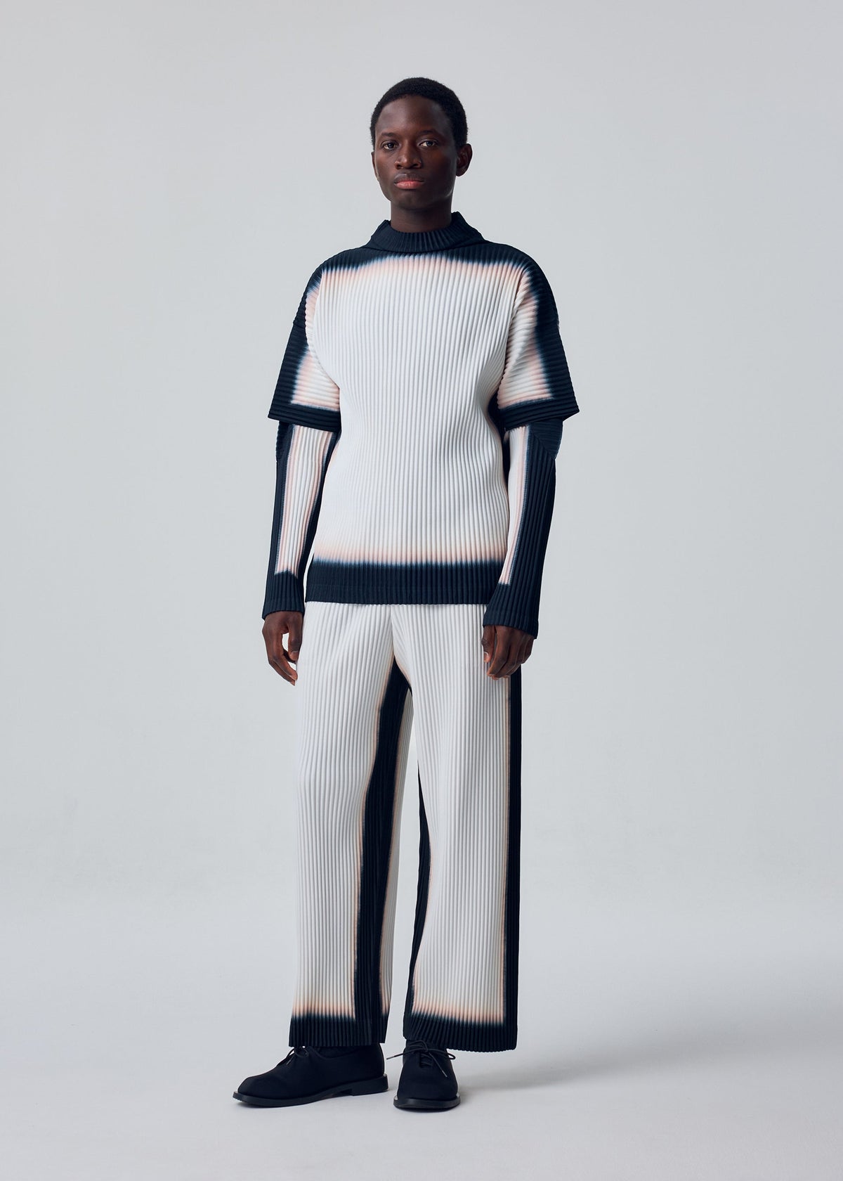 Zoomed view A person stands against a plain background, wearing the FRAMEWORK : CHUSEN set by HOMME PLISSÉ ISSEY MIYAKE—a pleated chusen dye top and matching pants with a white and black gradient, plus black shoes.
