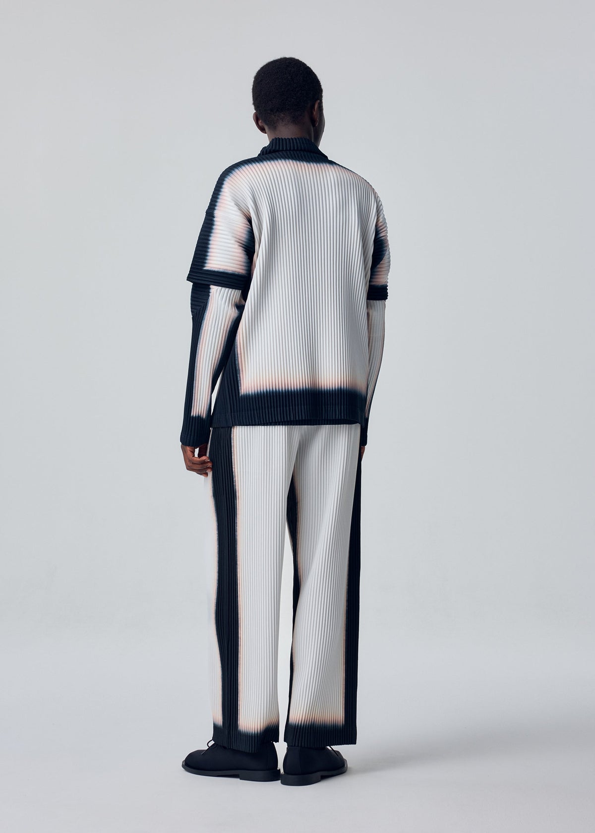 A person stands facing away, dressed in HOMME PLISSÉ ISSEY MIYAKE’s FRAMEWORK : CHUSEN—pleated, gradient attire with layered sleeves and matching wide-leg pants in black, white, and beige against a light background. view 5 of 6