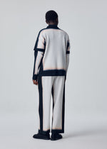 A person stands facing away, dressed in HOMME PLISSÉ ISSEY MIYAKE’s FRAMEWORK : CHUSEN—pleated, gradient attire with layered sleeves and matching wide-leg pants in black, white, and beige against a light background.