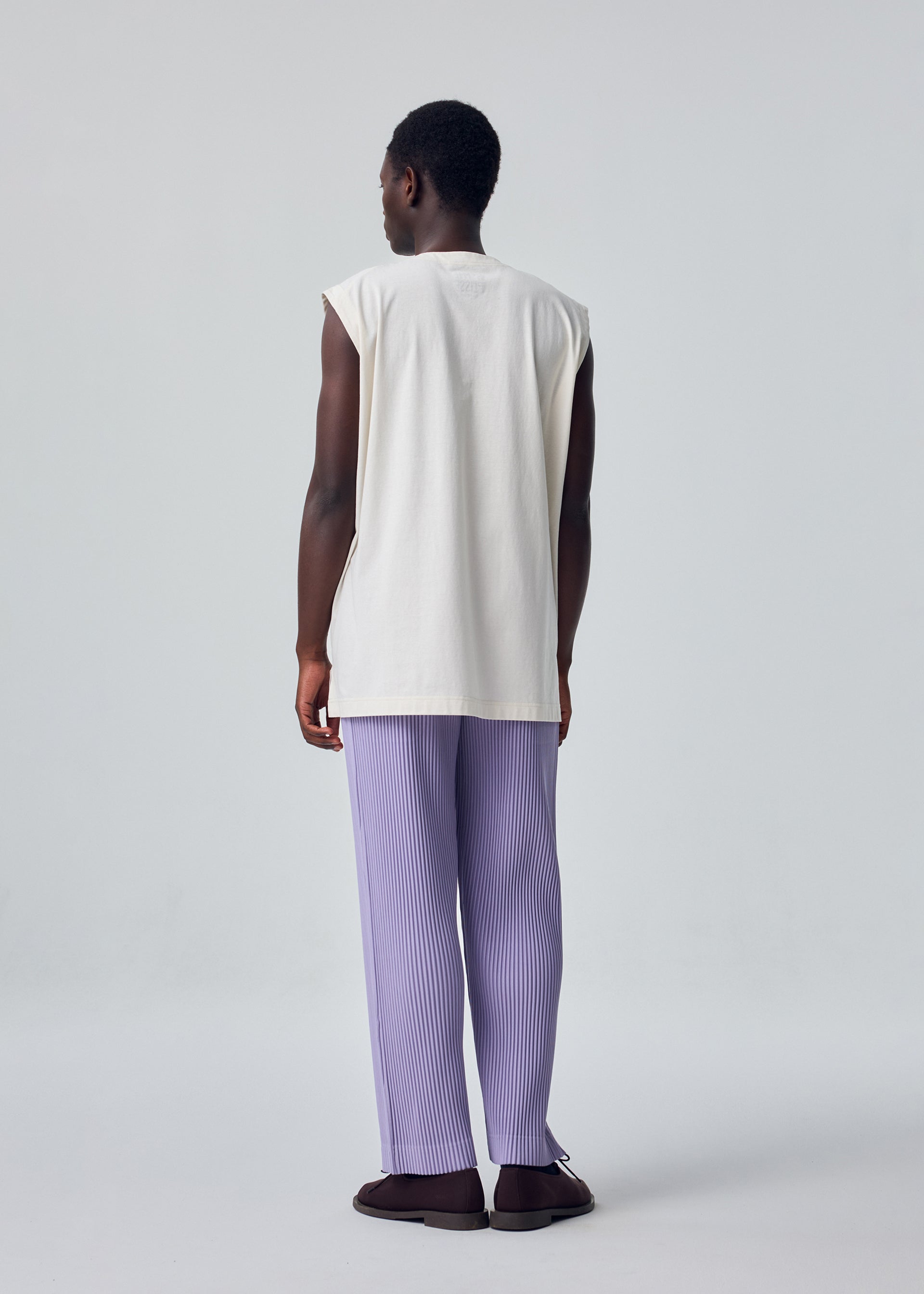 Zoomed view A person stands facing away from the camera in a HOMME PLISSÉ ISSEY MIYAKE HARD TWIST JERSEY sleeveless round-neck top, loose lavender pleated pants, and dark shoes against a plain light gray background.