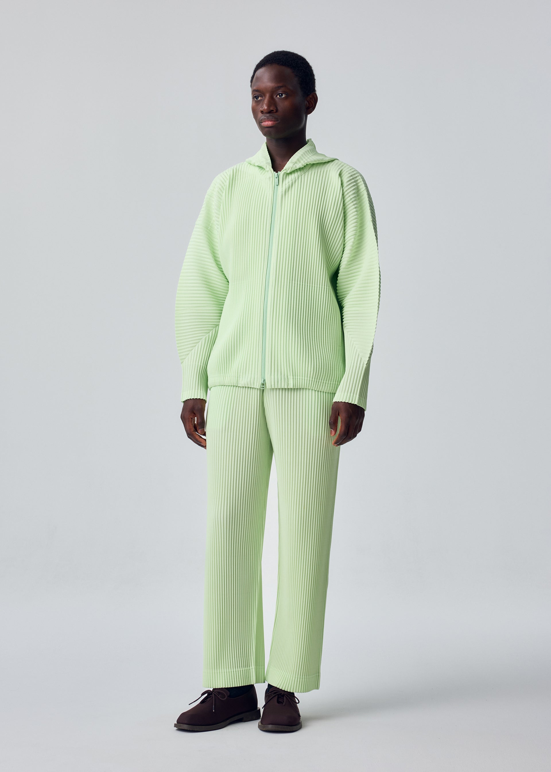 Zoomed view A person stands facing forward in HOMME PLISSÉ ISSEY MIYAKE's MONTHLY COLORS : JULY—light green, quick-drying polyester zip-up hoodie and matching wide-leg pants—and dark brown shoes, set against a plain light background.