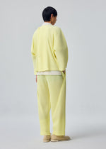 A person with short dark hair stands facing away, wearing the SLASH ensemble by HOMME PLISSÉ ISSEY MIYAKE—a loose, pale yellow outfit featuring diagonal pleats and matching pants with slash pockets—paired with beige shoes against a plain light background.