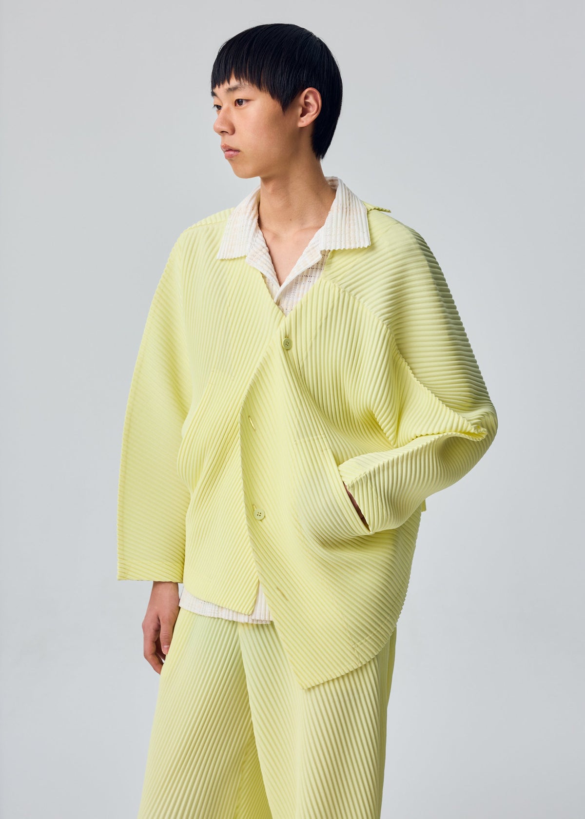 A person with short black hair wears the SLASH ensemble by HOMME PLISSÉ ISSEY MIYAKE—comprising a light yellow, pleated jacket with distinctive diagonal pleats and matching pants—layered over a white shirt, standing against a plain light gray background. view 7 of 7