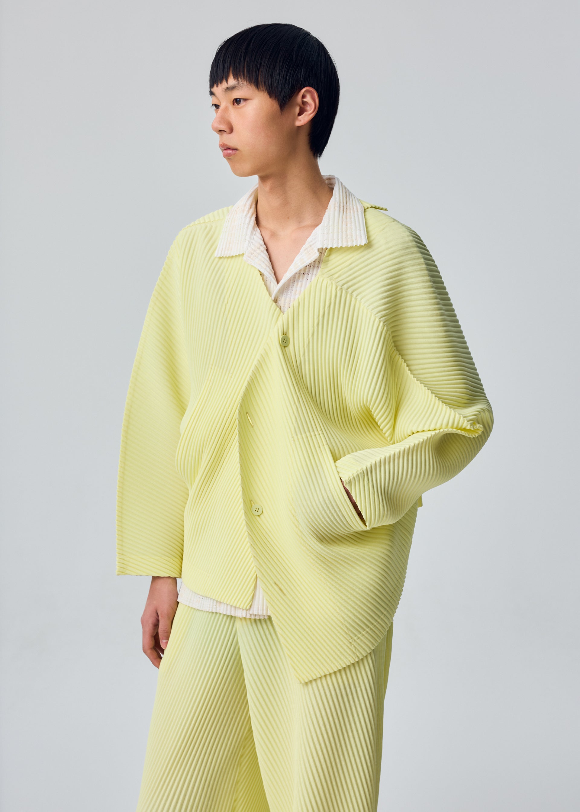 Zoomed view A person with short black hair wears the SLASH ensemble by HOMME PLISSÉ ISSEY MIYAKE—comprising a light yellow, pleated jacket with distinctive diagonal pleats and matching pants—layered over a white shirt, standing against a plain light gray background.
