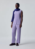 A person stands against a plain light background, wearing HOMME PLISSÉ ISSEY MIYAKE's MONTHLY COLORS : JULY—lavender sleeveless top and matching pants—plus a textured purple scarf over one shoulder. They have short hair and a slight smile.