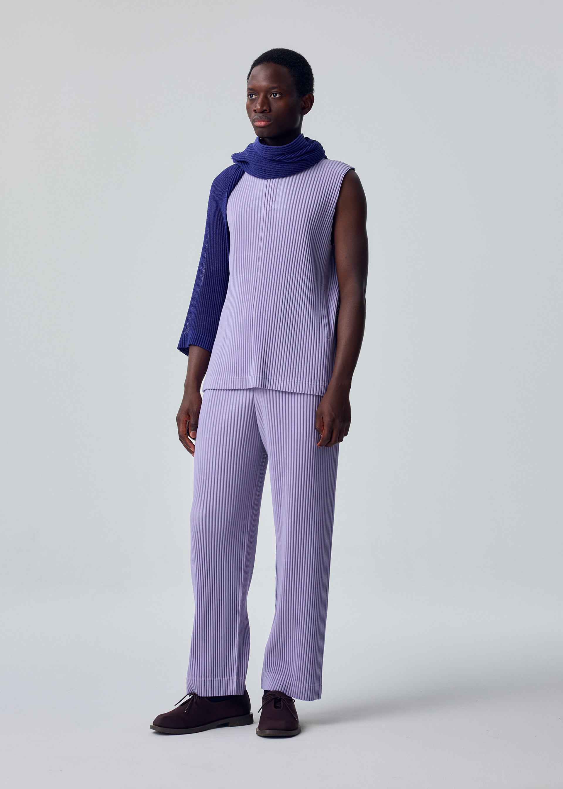 Zoomed view A person stands against a plain light background, wearing HOMME PLISSÉ ISSEY MIYAKE's MONTHLY COLORS : JULY—lavender sleeveless top and matching pants—plus a textured purple scarf over one shoulder. They have short hair and a slight smile.
