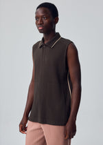 A person wearing the EMBROIDERY STRIPES dark brown sleeveless, ribbed collared top with a button at the neck from HOMME PLISSÉ ISSEY MIYAKE and pink pleated pants stands against a plain light gray background.