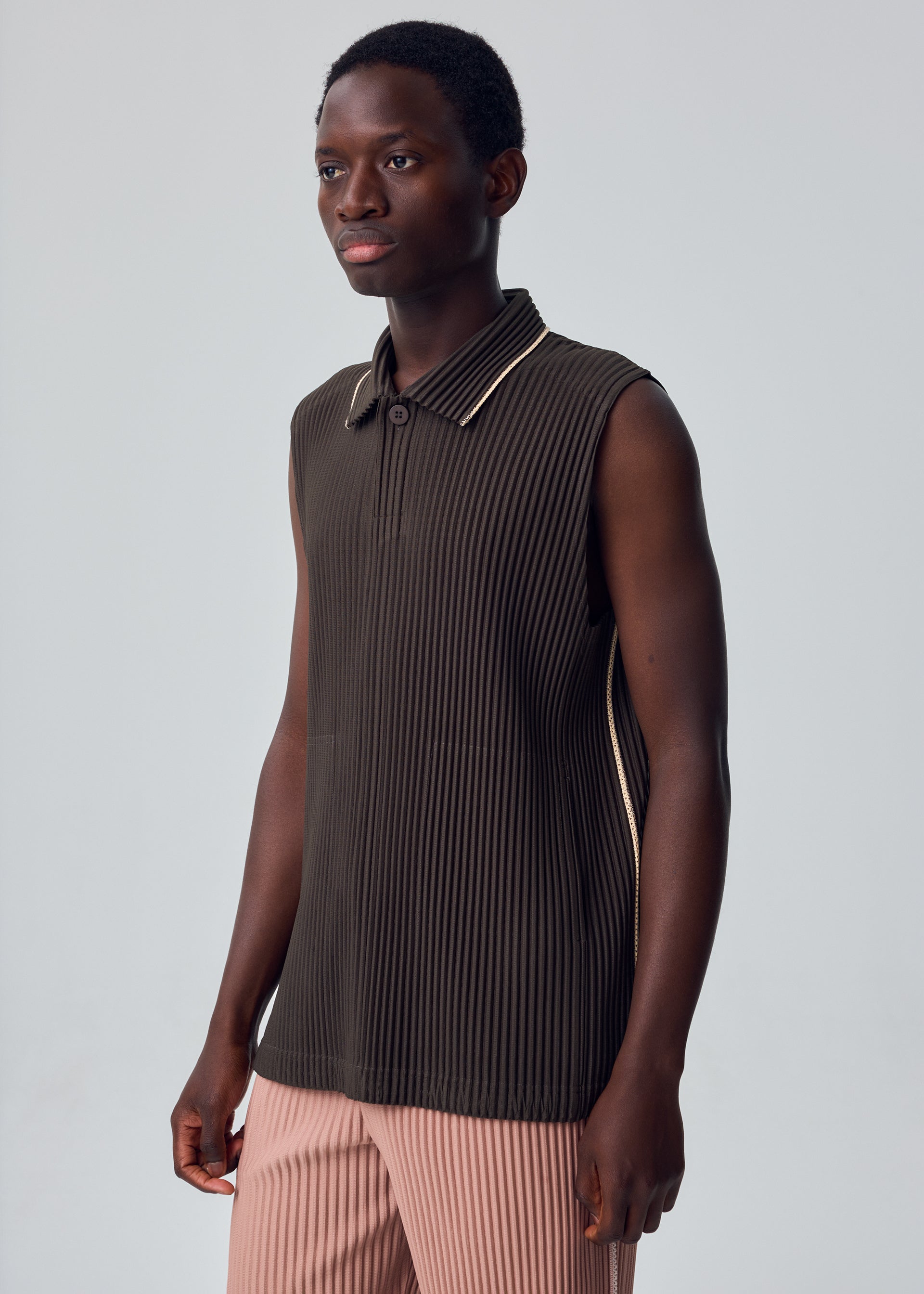 Zoomed view A person wearing the EMBROIDERY STRIPES dark brown sleeveless, ribbed collared top with a button at the neck from HOMME PLISSÉ ISSEY MIYAKE and pink pleated pants stands against a plain light gray background.