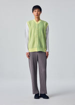 A person faces forward, wearing the IN THE SHADOW KNIT by HOMME PLISSÉ ISSEY MIYAKE over a white long-sleeve shirt, paired with gray pleated pants and black shoes against a plain light background.