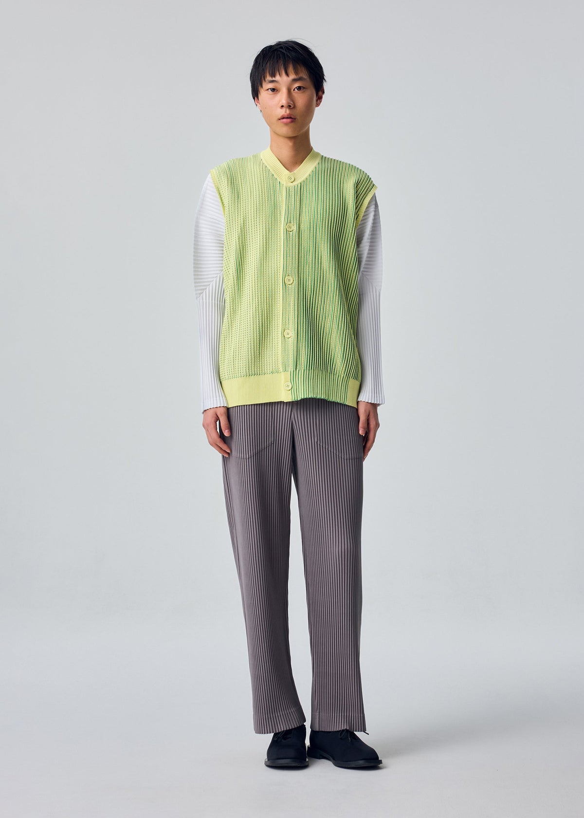 Zoomed view A person faces forward, wearing the IN THE SHADOW KNIT by HOMME PLISSÉ ISSEY MIYAKE over a white long-sleeve shirt, paired with gray pleated pants and black shoes against a plain light background.