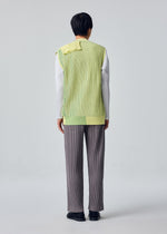 A person faces away in gray pants, black shoes, and the HOMME PLISSÉ ISSEY MIYAKE IN THE SHADOW KNIT— a long, ribbed, yellow-green vest over a white shirt— against a plain gray background.