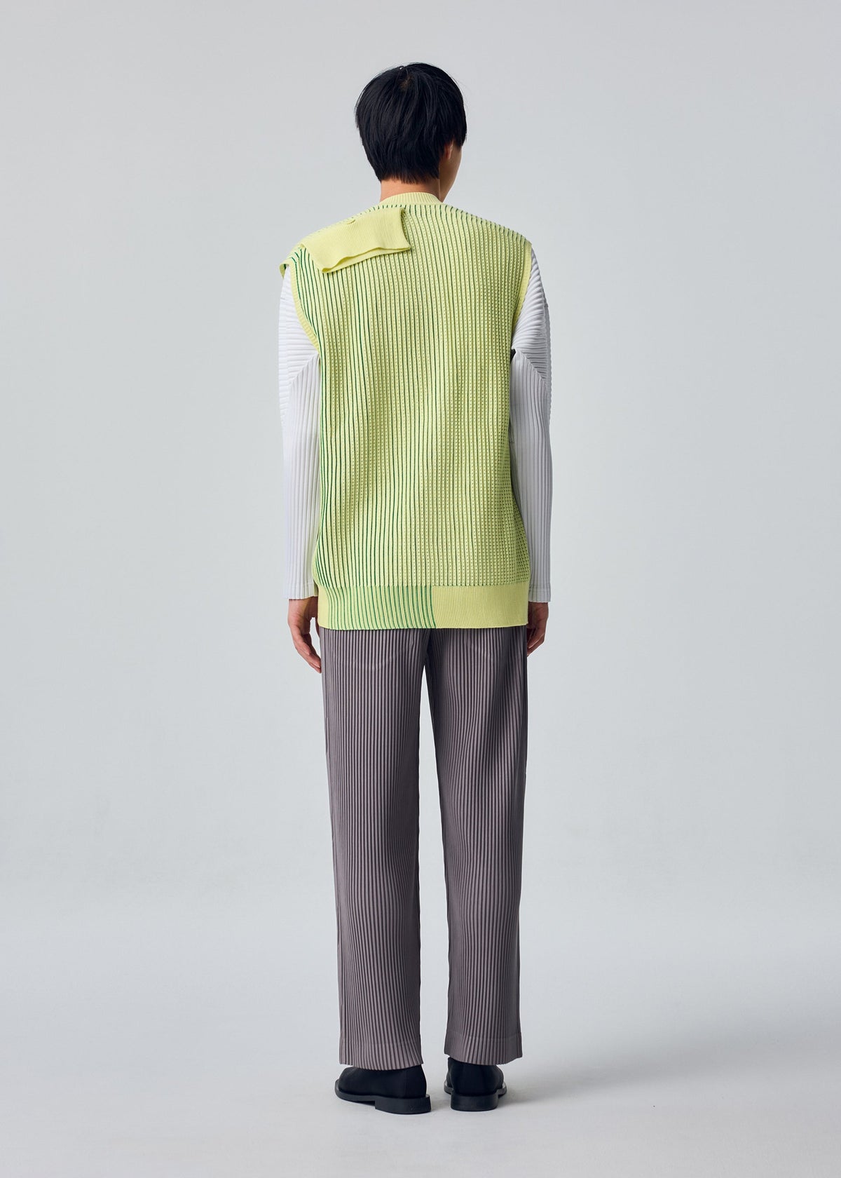Zoomed view A person faces away in gray pants, black shoes, and the HOMME PLISSÉ ISSEY MIYAKE IN THE SHADOW KNIT— a long, ribbed, yellow-green vest over a white shirt— against a plain gray background.
