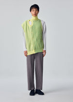 A person stands facing forward in the IN THE SHADOW KNIT by HOMME PLISSÉ ISSEY MIYAKE, layered over a white ribbed top and loose gray pinstriped pants against a light gray background.