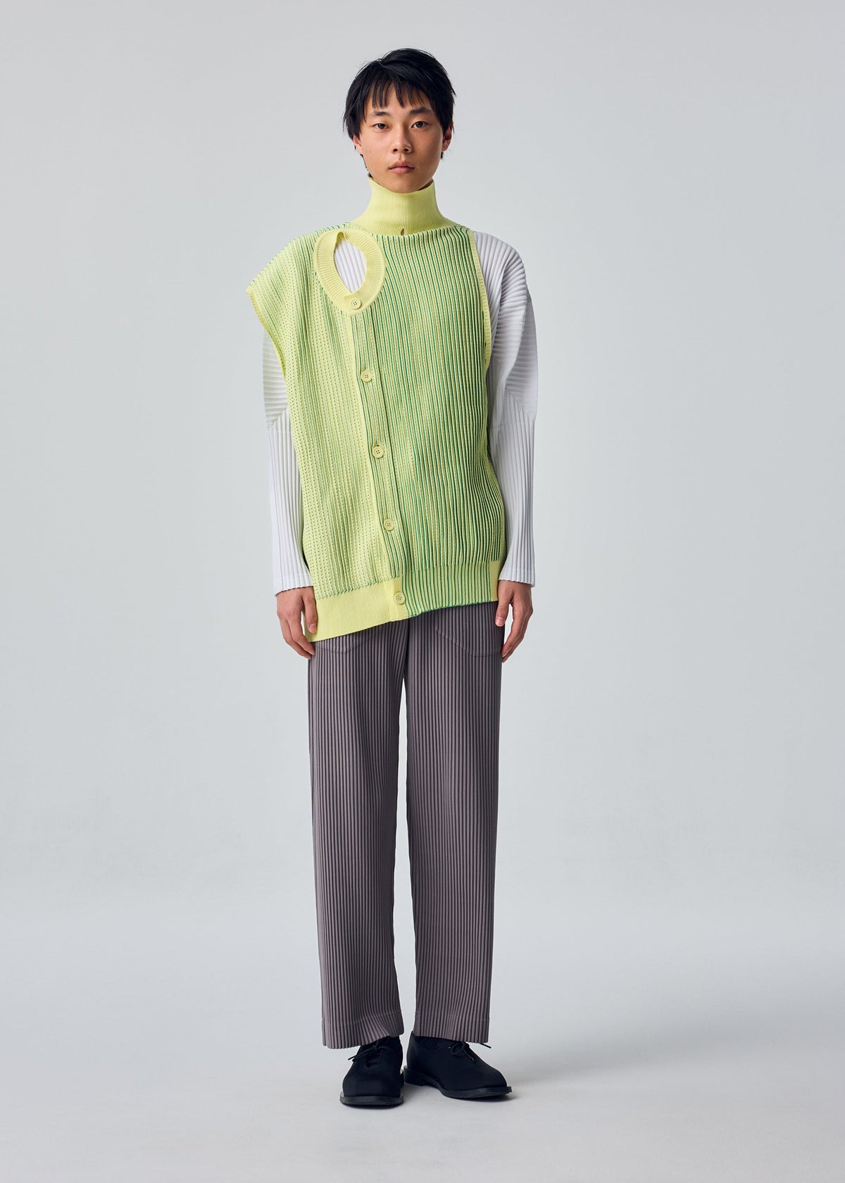 Zoomed view A person stands facing forward in the IN THE SHADOW KNIT by HOMME PLISSÉ ISSEY MIYAKE, layered over a white ribbed top and loose gray pinstriped pants against a light gray background.