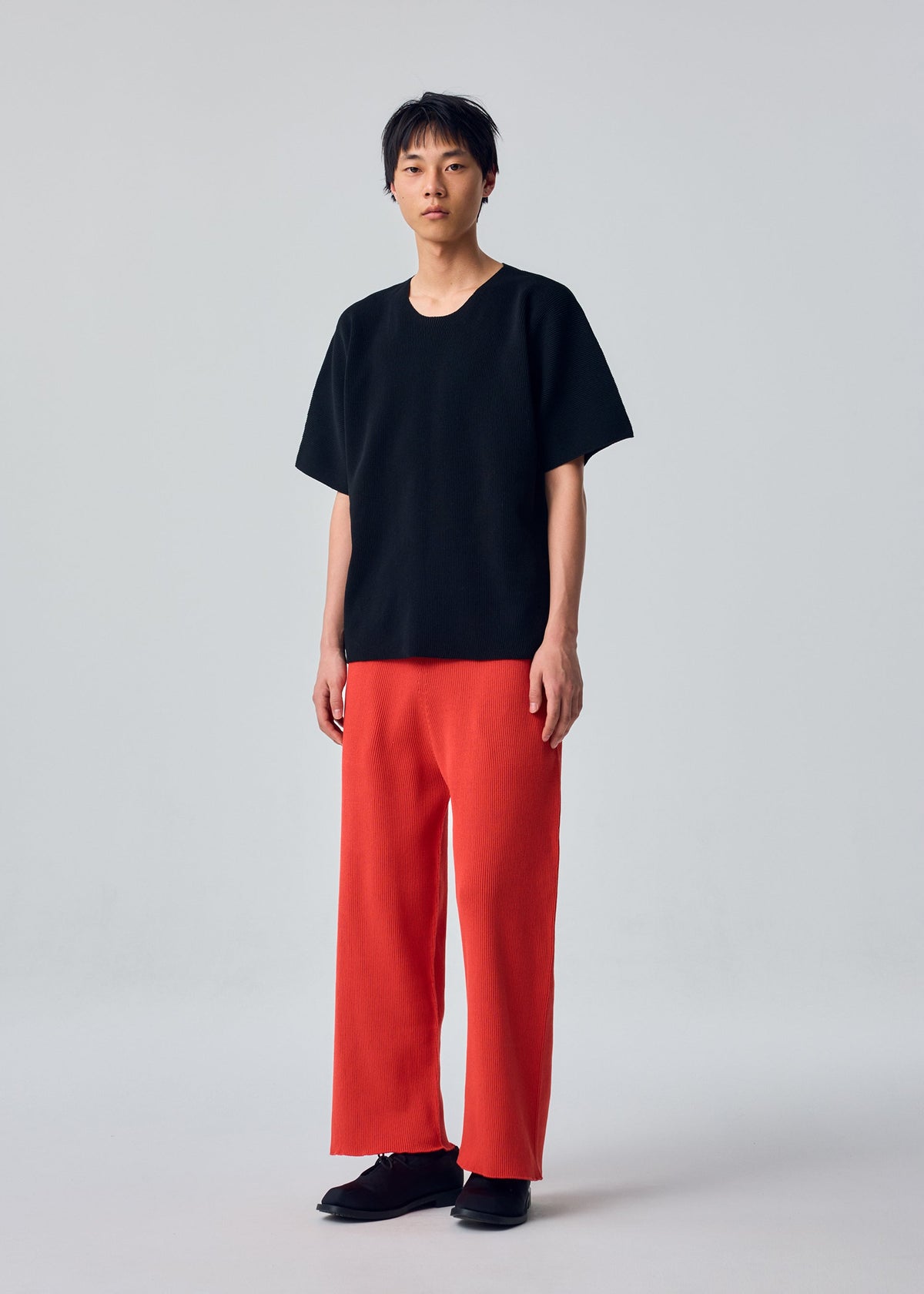 A person stands against a plain background wearing the HOMME PLISSÉ ISSEY MIYAKE BASIC KNIT, paired with wide red pleated jersey pants and black shoes. view 4 of 5