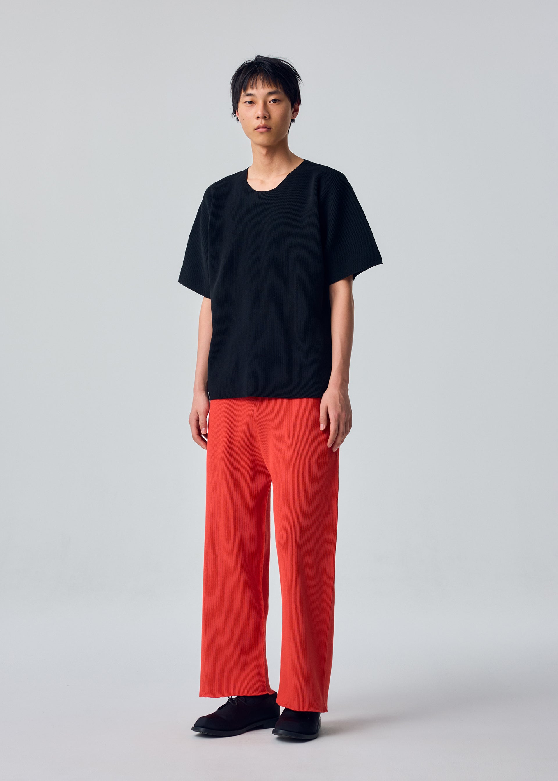 Zoomed view A person stands against a plain background wearing the HOMME PLISSÉ ISSEY MIYAKE BASIC KNIT, paired with wide red pleated jersey pants and black shoes.