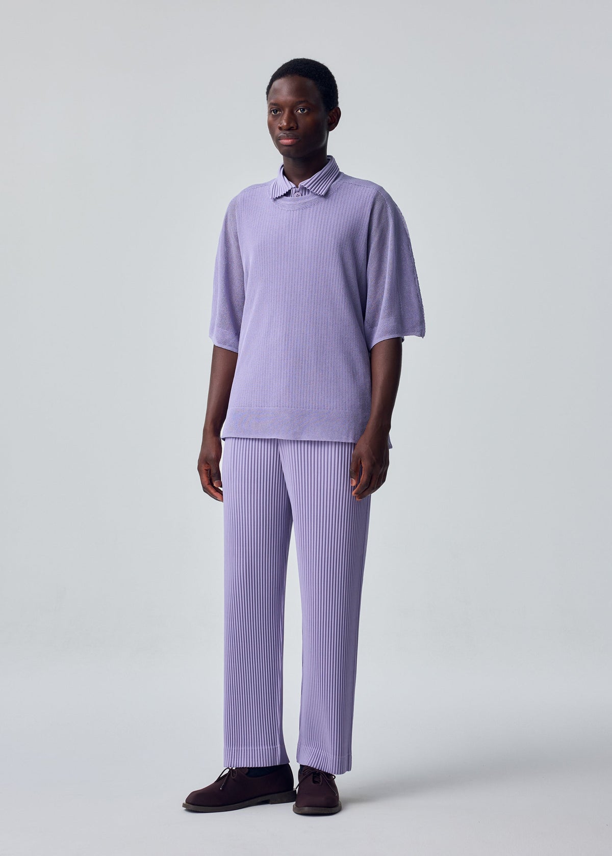 A person stands against a light gray backdrop in HOMME PLISSÉ ISSEY MIYAKE's WASHI FRAMEWORK-T, a loose lilac short-sleeved polo layered over a round-neck shirt and matching pleated pants, with dark shoes, arms relaxed and gazing to the side. view 5 of 6