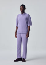 A person stands against a light gray backdrop in HOMME PLISSÉ ISSEY MIYAKE's WASHI FRAMEWORK-T, a loose lilac short-sleeved polo layered over a round-neck shirt and matching pleated pants, with dark shoes, arms relaxed and gazing to the side.