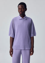 A person with short hair and a neutral expression stands against a plain light gray background, wearing the HOMME PLISSÉ ISSEY MIYAKE WASHI FRAMEWORK-T in light purple with elbow-length sleeves and matching ribbed pants.