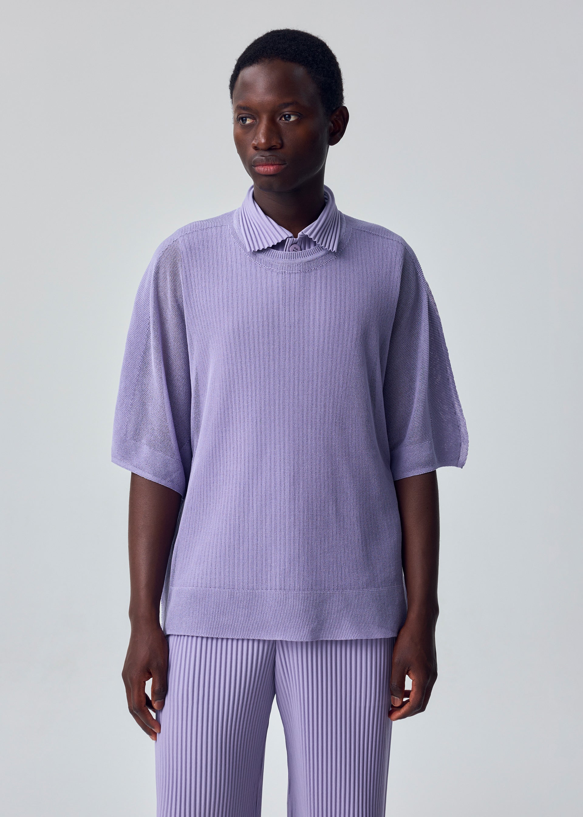 Zoomed view A person with short hair and a neutral expression stands against a plain light gray background, wearing the HOMME PLISSÉ ISSEY MIYAKE WASHI FRAMEWORK-T in light purple with elbow-length sleeves and matching ribbed pants.