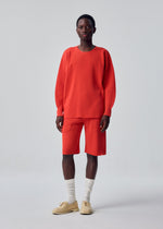 A person stands against a plain background wearing the HOMME PLISSÉ ISSEY MIYAKE BASIC KNIT: a loose-fitting red pleated jersey top paired with matching red knee-length shorts, cream socks, and beige shoes.