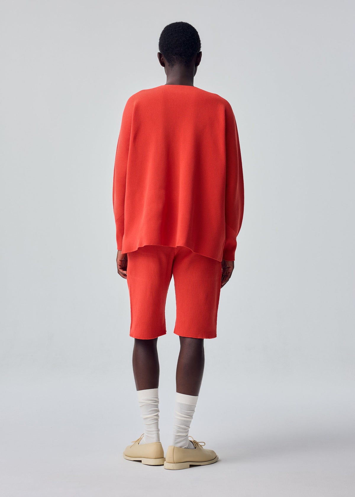 A person stands facing away, wearing the HOMME PLISSÉ ISSEY MIYAKE BASIC KNIT—a loose-fitting, bright red pleated jersey top paired with matching knee-length shorts crafted from regenerated thread—styled with white socks and beige shoes against a plain light background. view 5 of 5