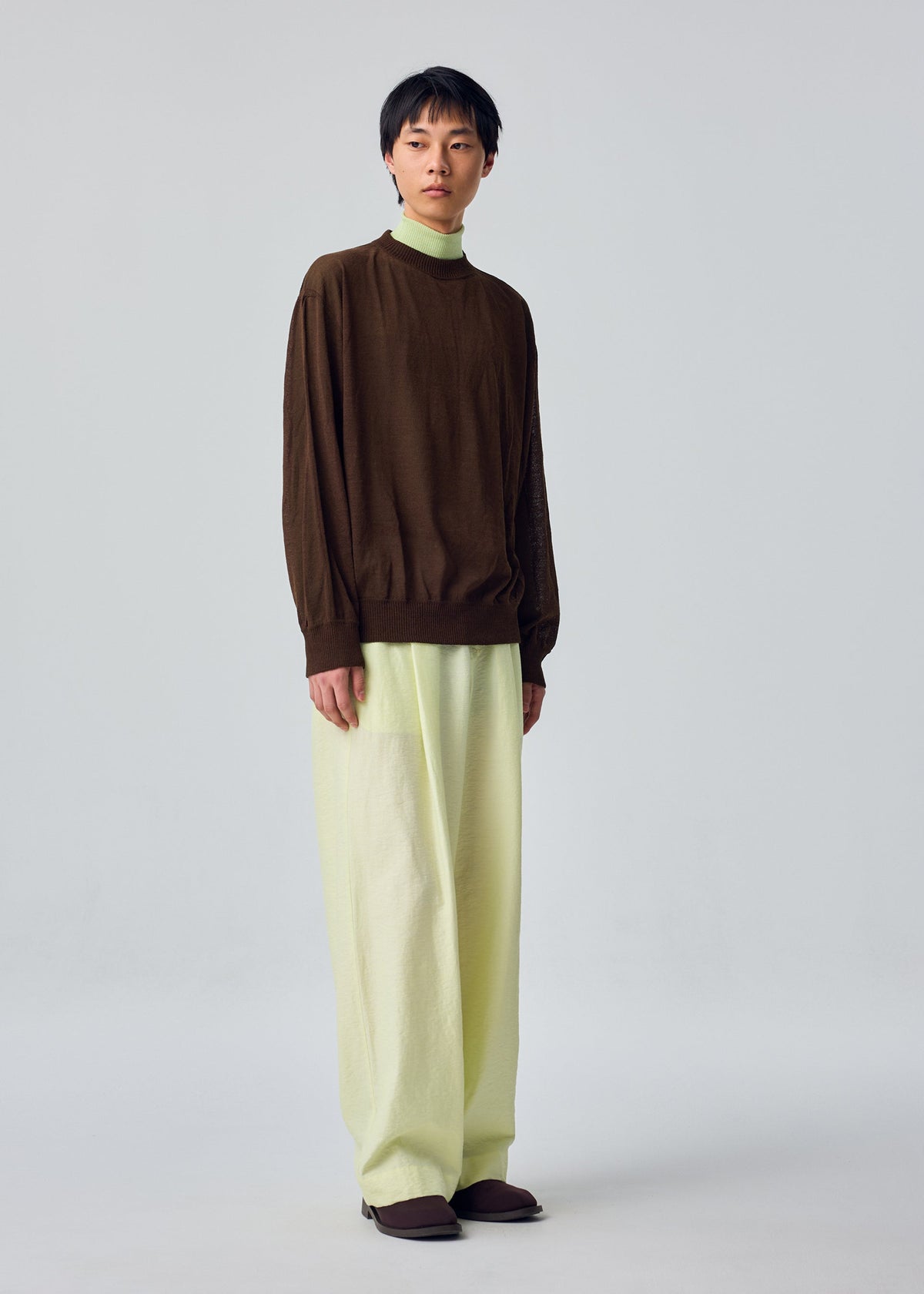 A person stands against a plain background wearing the KNIT UNRAVELED long-sleeve brown top by HOMME PLISSÉ ISSEY MIYAKE layered over a light green turtleneck, paired with wide-legged light green pants and brown shoes for a contemporary, layered look. view 6 of 7
