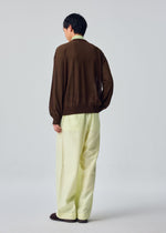 A person stands facing away, wearing the HOMME PLISSÉ ISSEY MIYAKE KNIT UNRAVELED top in brown, paired with light yellow wide-leg pants and brown shoes, against a plain light gray background.