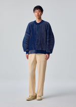 A person faces forward in the IN THE SHADOW KNIT by HOMME PLISSÉ ISSEY MIYAKE, styled with cream pleated pants and beige shoes against a plain light background.