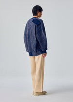 A person faces away, styled in IN THE SHADOW KNIT by HOMME PLISSÉ ISSEY MIYAKE with a blue pinstriped jacket, cream wide-leg pants, and beige shoes against a plain light background.
