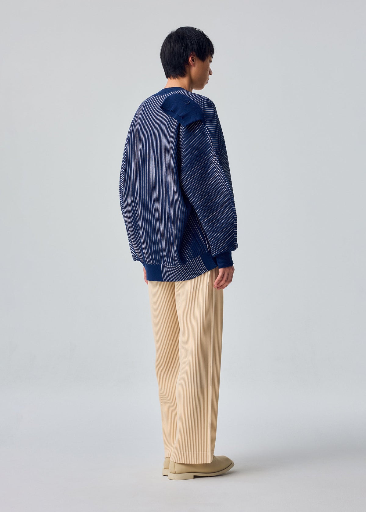 Zoomed view A person faces away, styled in IN THE SHADOW KNIT by HOMME PLISSÉ ISSEY MIYAKE with a blue pinstriped jacket, cream wide-leg pants, and beige shoes against a plain light background.