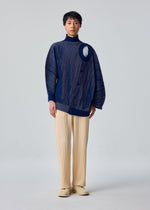 A person stands in a studio wearing the IN THE SHADOW KNIT by HOMME PLISSÉ ISSEY MIYAKE, a blue long-sleeve top with an asymmetrical cutout, paired with beige pleated pants against a plain light background.