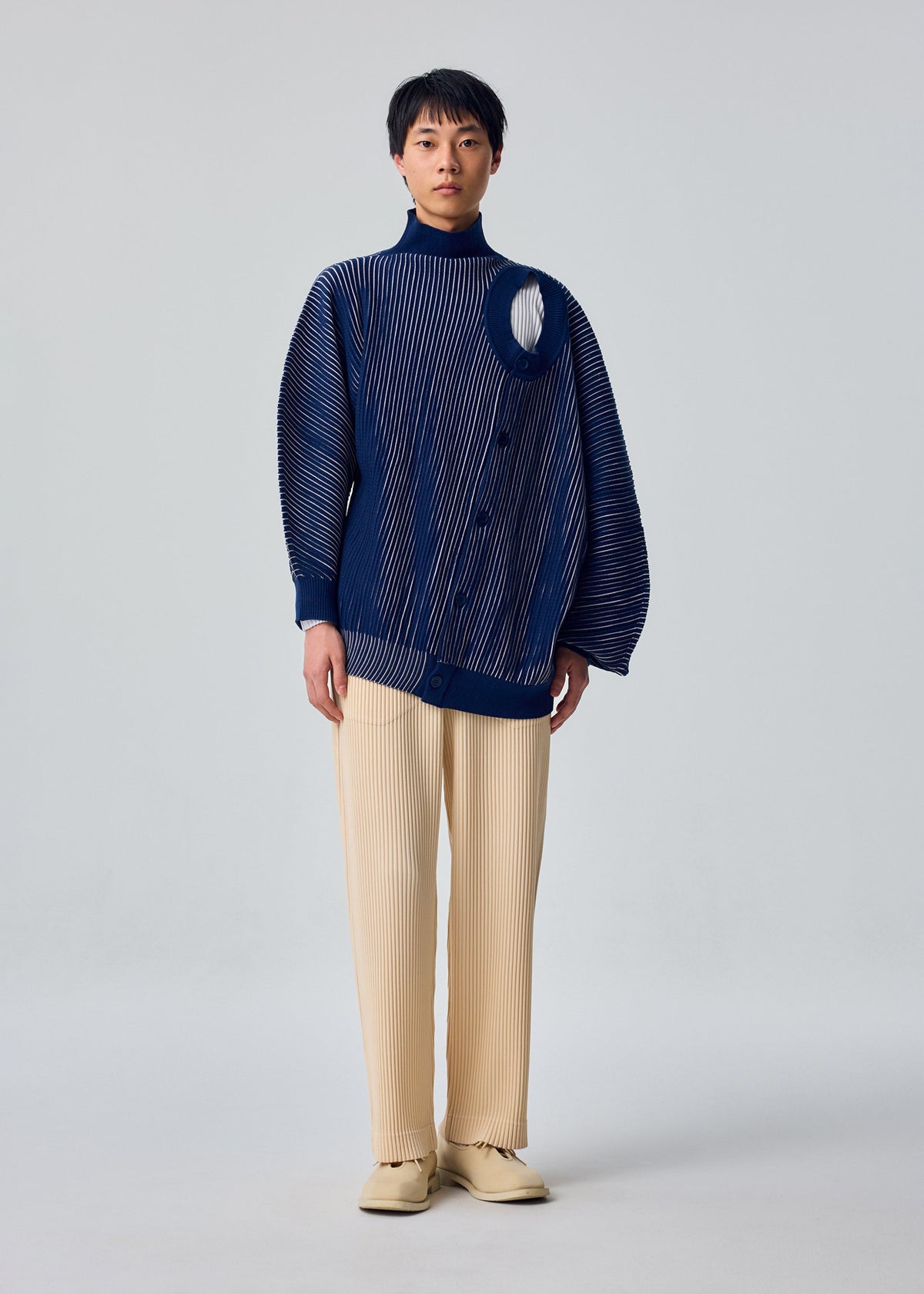 Zoomed view A person stands in a studio wearing the IN THE SHADOW KNIT by HOMME PLISSÉ ISSEY MIYAKE, a blue long-sleeve top with an asymmetrical cutout, paired with beige pleated pants against a plain light background.