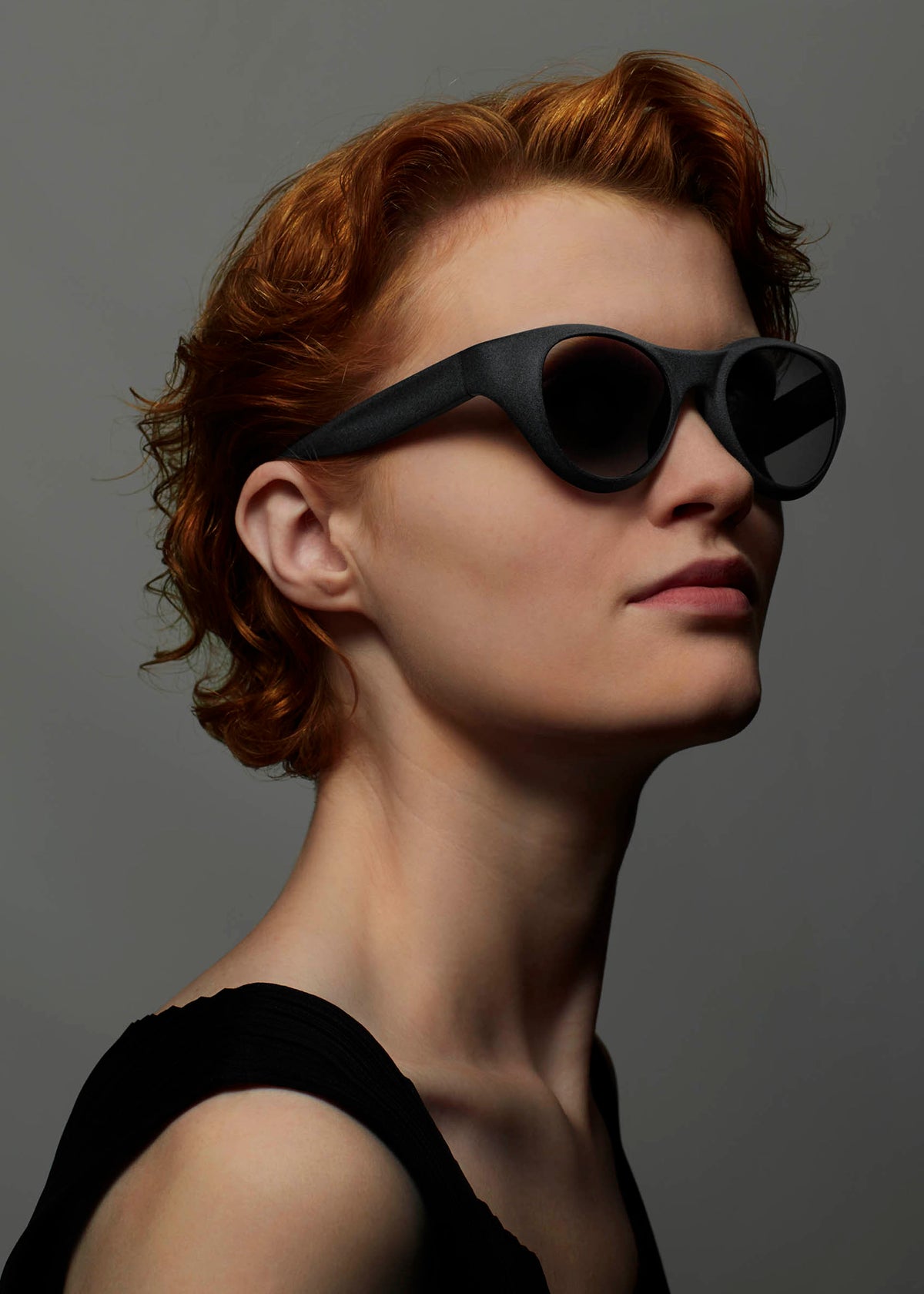 A person with short, wavy red hair wears ISSEY MIYAKE EYES GRIT-BOSTON sunglasses and a black sleeveless top, posing confidently against a plain gray background. view 6 of 6