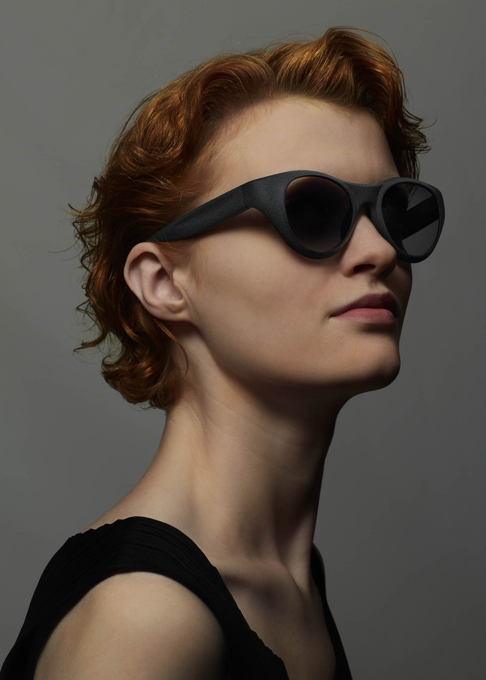 Zoomed view A person with short, wavy red hair wears ISSEY MIYAKE EYES GRIT-BOSTON sunglasses and a black sleeveless top, posing confidently against a plain gray background.