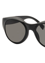 Close-up of the ISSEY MIYAKE EYES GRIT-BOSTON sunglasses in black, featuring an oval shape and dark lenses, set against a white background. Only part of the frame and one lens are fully visible.