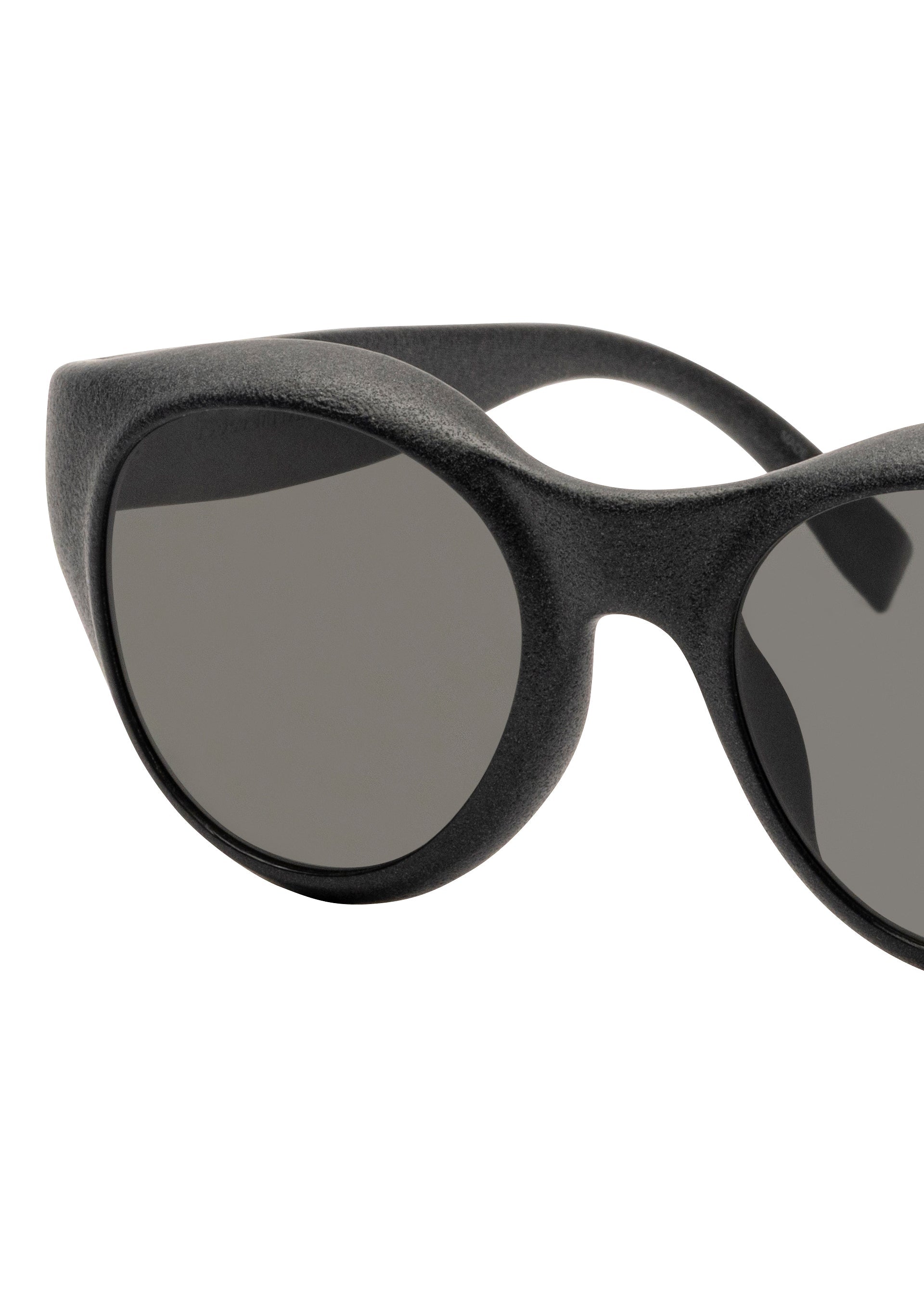 Zoomed view Close-up of the ISSEY MIYAKE EYES GRIT-BOSTON sunglasses in black, featuring an oval shape and dark lenses, set against a white background. Only part of the frame and one lens are fully visible.