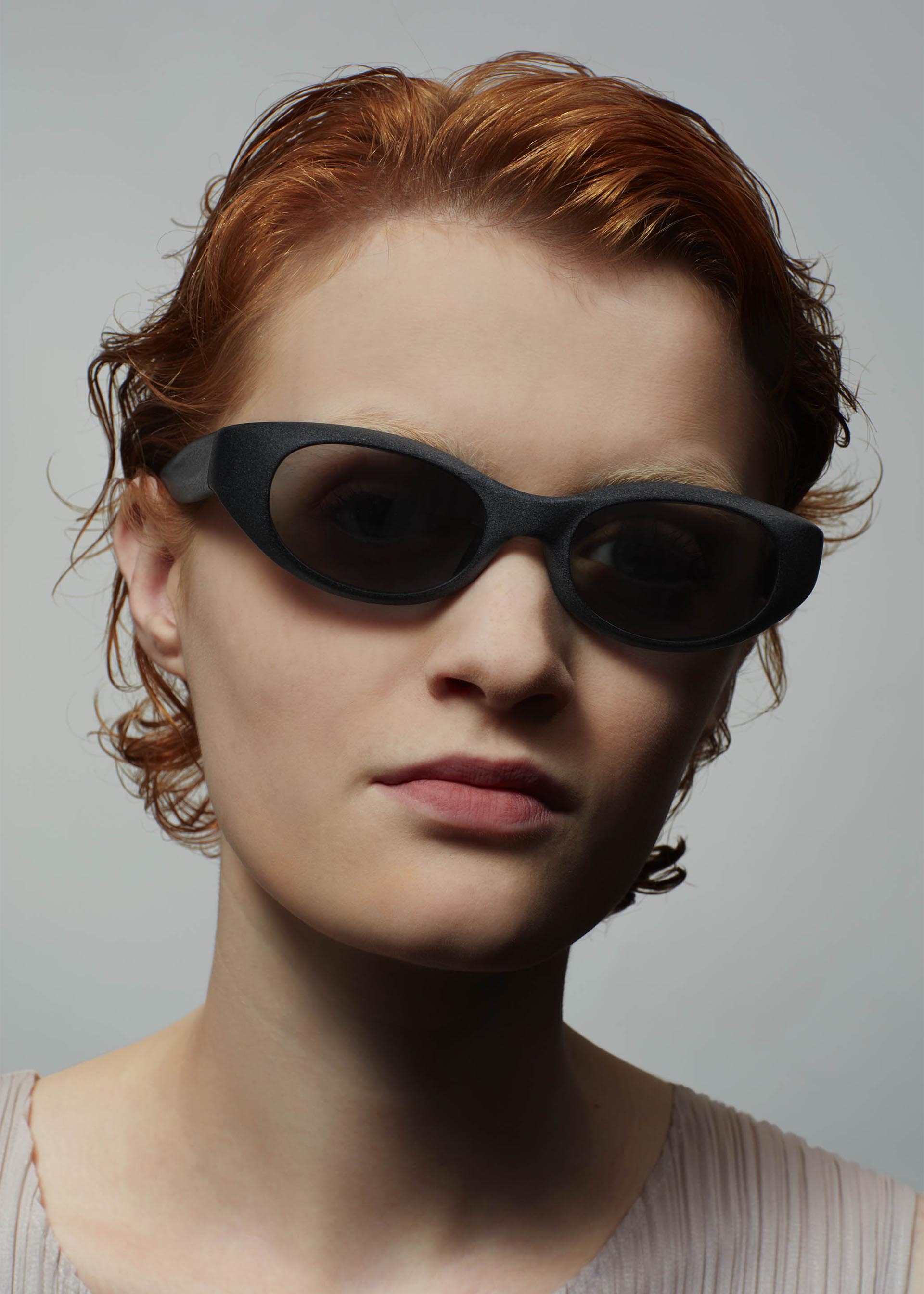 Zoomed view A person with short, wavy red hair wears ISSEY MIYAKE EYES GRIT-SQUARE black 3D printed sunglasses and a light-colored top, facing the camera against a plain gray background.