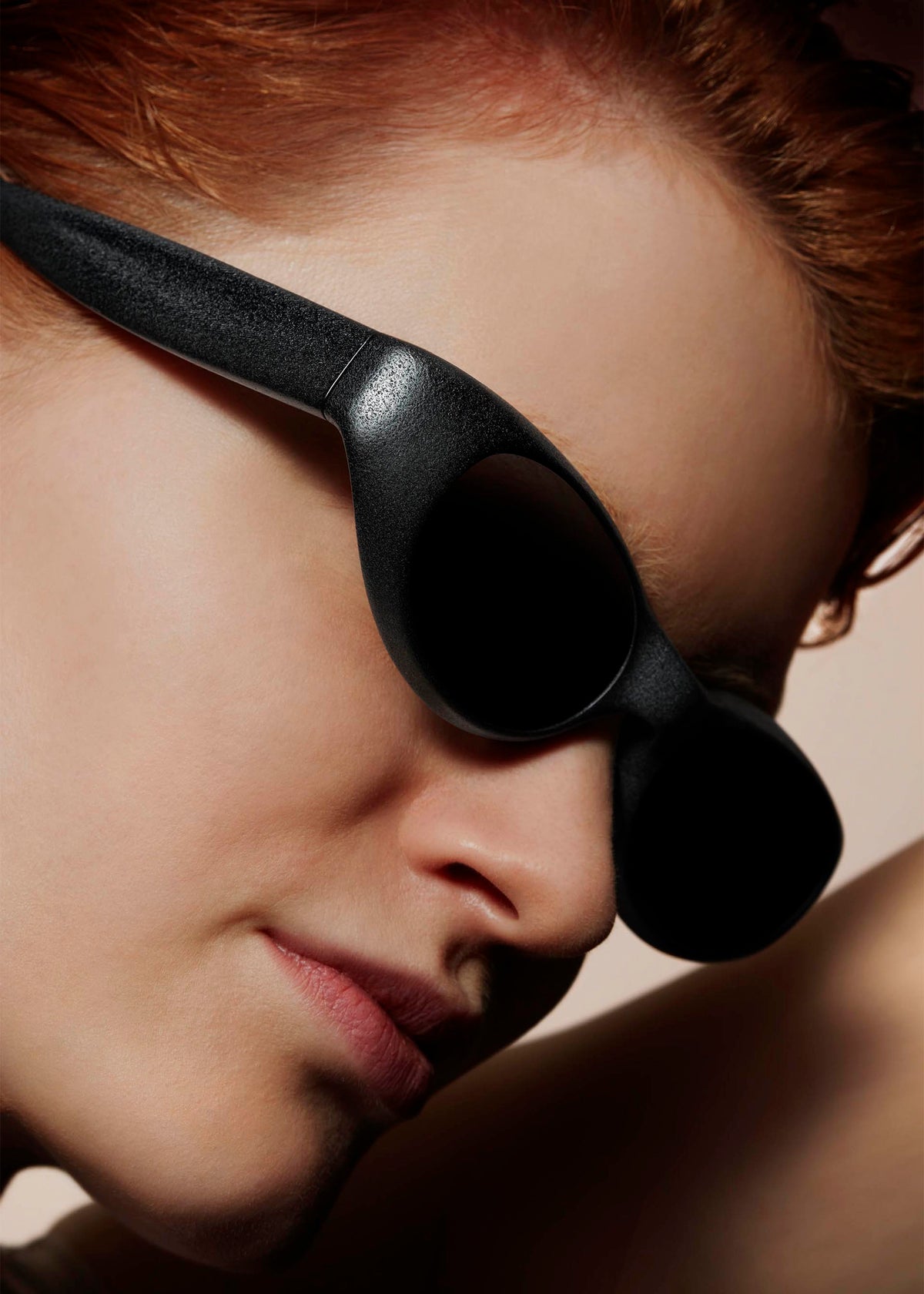 A person with fair skin and short, reddish hair wears large GRIT-SQUARE sunglasses by ISSEY MIYAKE EYES. The black, 3D printed frames stand out as smooth skin and facial features are highlighted against a neutral backdrop. view 7 of 7
