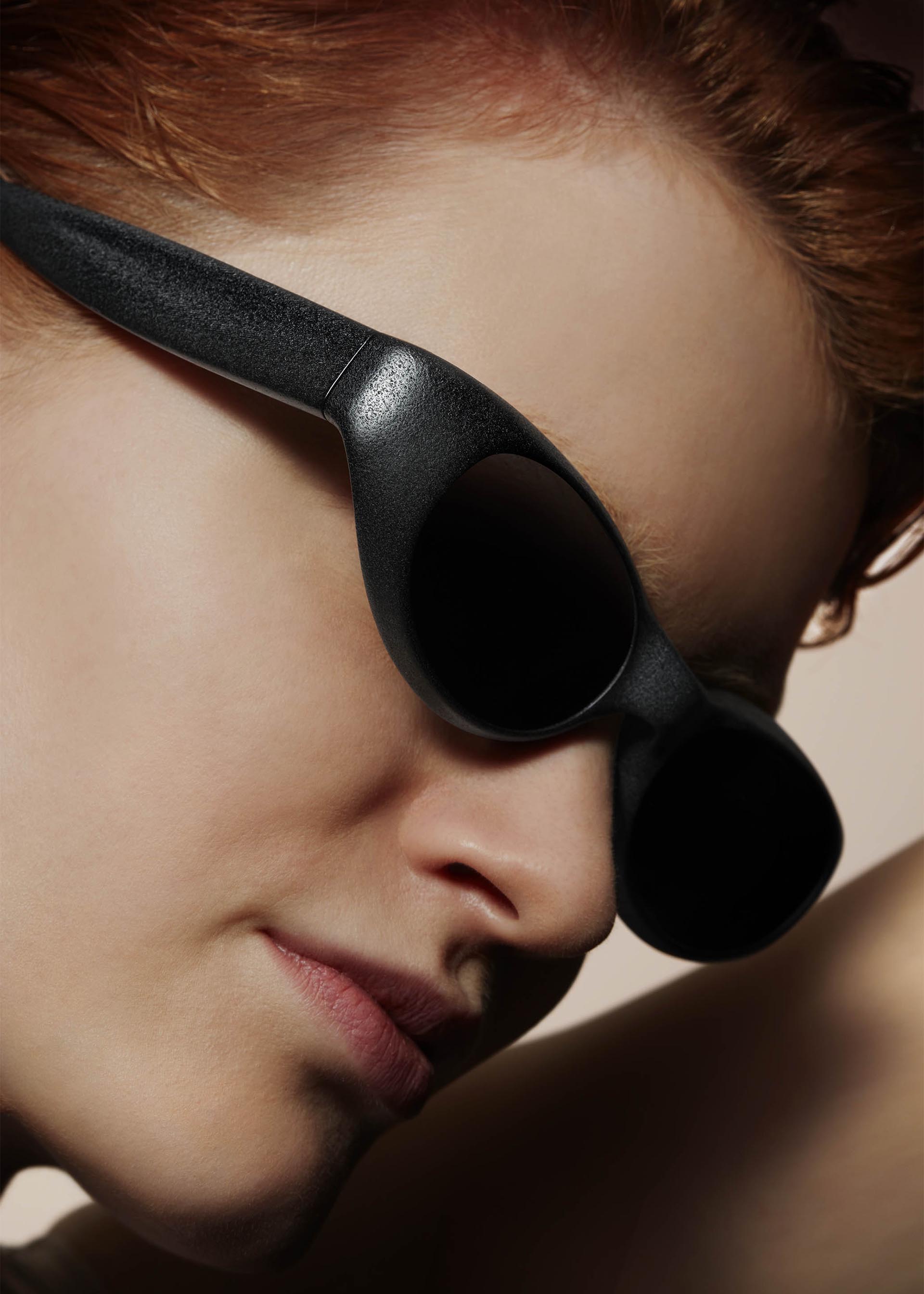 Zoomed view A person with fair skin and short, reddish hair wears large GRIT-SQUARE sunglasses by ISSEY MIYAKE EYES. The black, 3D printed frames stand out as smooth skin and facial features are highlighted against a neutral backdrop.